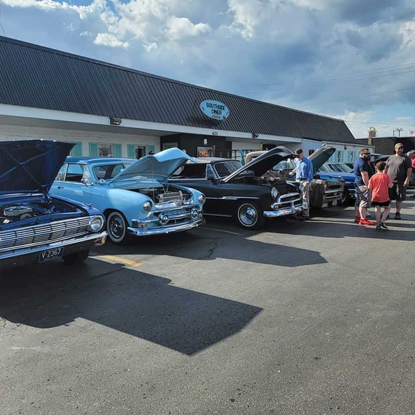 Classic cars with open hoods line up in front of the Southside Diner, with people admiring them.