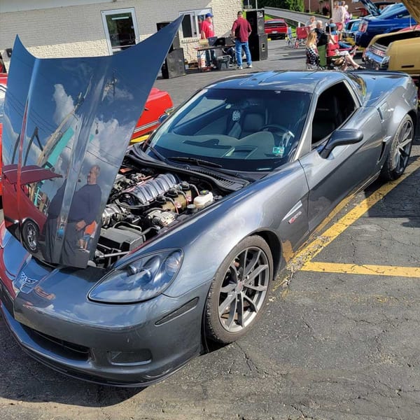 Gray Corvette with its hood open, parked in a lot with people and other cars in the background.