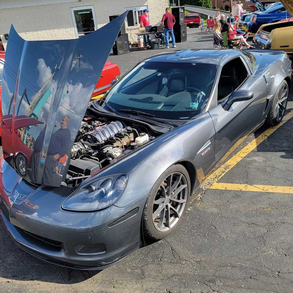 Gray Corvette with its hood open, parked in a lot with people and other cars in the background.